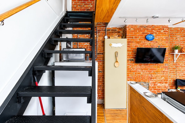 A kitchen with a brick wall, a microwave, a wooden spoon, and a television mounted on the wall.