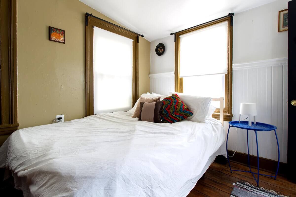 A bedroom with a bed, a small table with a lamp, and two windows with wooden frames.