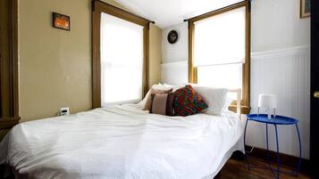 A bedroom with a bed, a small table with a lamp, and two windows with wooden frames.