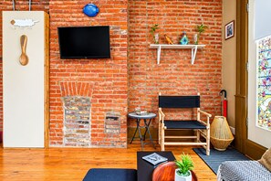 Une pièce avec un mur en briques, un téléviseur à écran plat fixé au-dessus d’une cheminée, une chaise en bois, une petite table ronde et une fenêtre offrant une vue colorée.