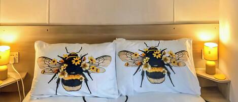 A bed with two pillows featuring a bee design, a bedside table with a lamp, and a wall-mounted artwork above the bed.