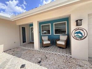 Une maison à l’extérieur bleu, avec deux chaises en osier, un panneau d’accueil et une terrasse carrelée.