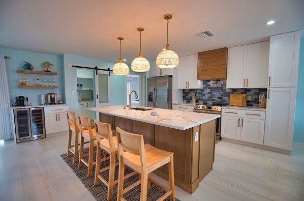 Una cocina moderna con una isla central, taburetes de bar, iluminación colgante y muebles blancos.