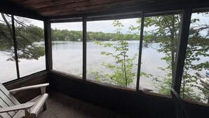 Un porche cubierto con vista a un lago, piso de madera y una sola silla Adirondack.
