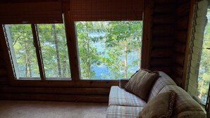 A log cabin interior with a plaid sofa and a view of a forest and lake through large windows. - Shady Rest Cabin #4, Sunset Views, Sandy Beach, Fishing, Trails, Firepit (Rhinelanderi)