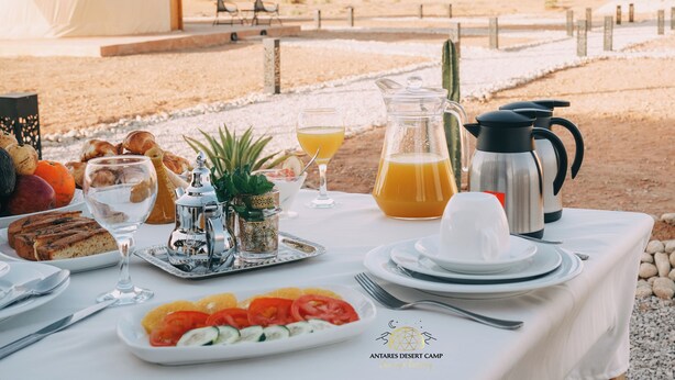 A desert camp breakfast table with a variety of foods, a pitcher of orange juice, and a view of the desert landscape. - Antares Desert Camp (Merzouga)