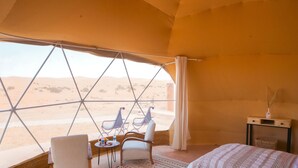 A tent with a view of the desert. Inside, there is a bed, a table with chairs, and a small side table with a vase. - Antares Desert Camp (Merzouga)