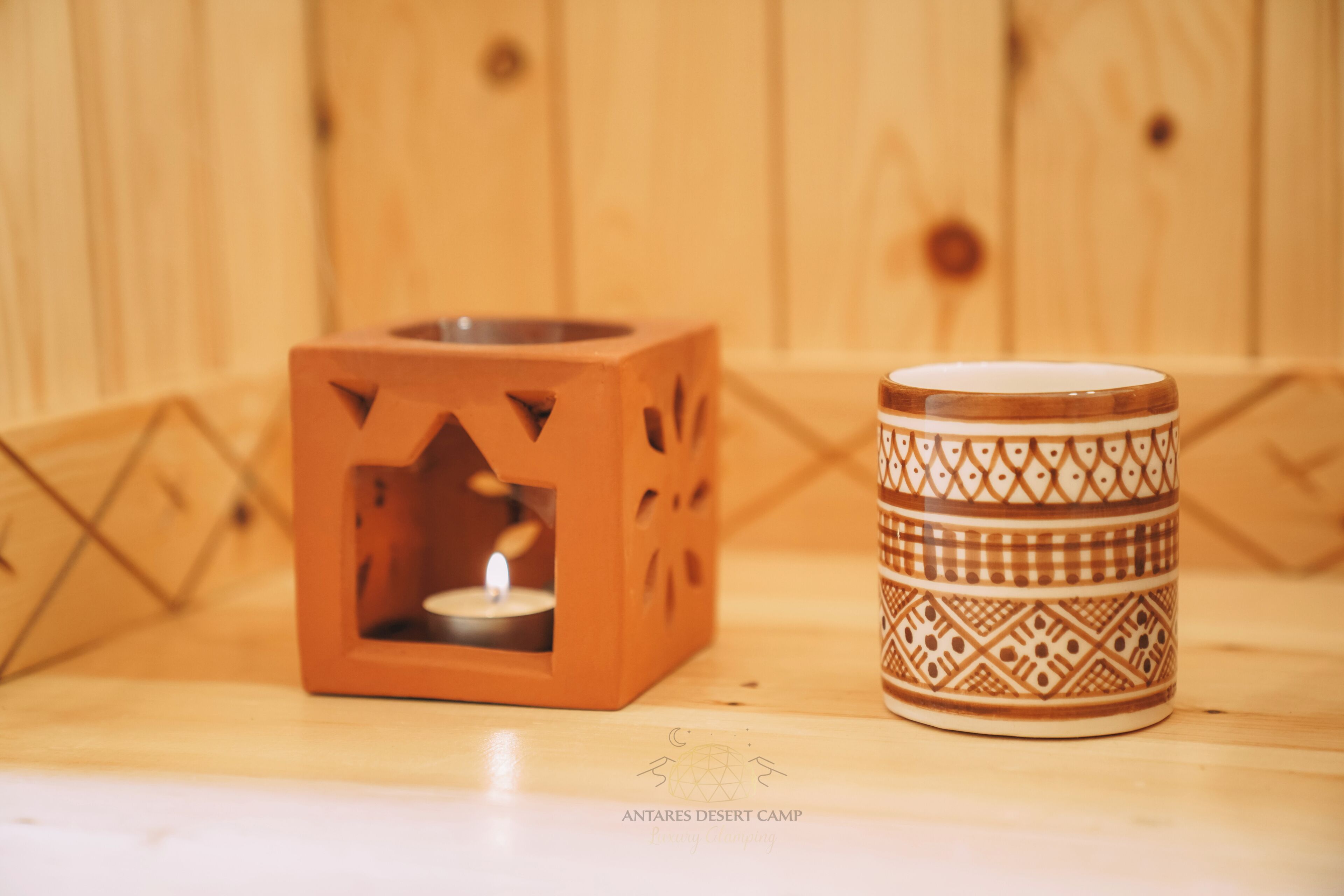 A wooden cube-shaped candle holder with a lit candle inside, next to a patterned ceramic mug on a wooden surface.