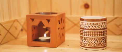 Un portalápices de madera con forma cúbica y una vela encendida en su interior, junto a una taza de cerámica estampada sobre una superficie de madera.