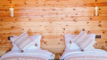 Interior de una cabaña de madera con dos camas, almohadones estampados y lámparas fijadas a la pared.
