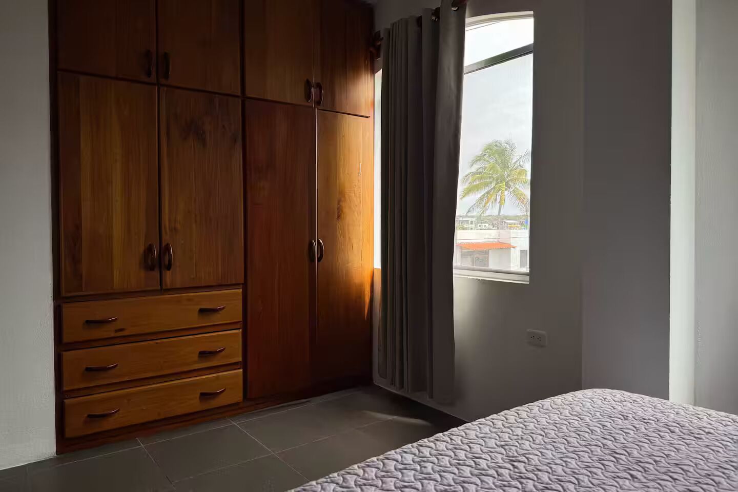 A bedroom with a large wooden wardrobe, a bed, and a window with a view of palm trees.