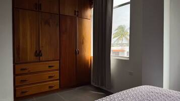 A bedroom with a large wooden wardrobe, a bed, and a window with a view of palm trees.