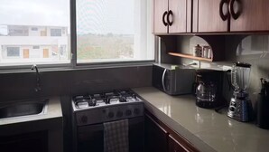 Een keuken met een gasfornuis, magnetron, koffiezetapparaat en blender. Er is een raam met uitzicht op gebouwen.