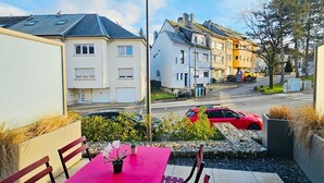 Une terrasse couverte avec une table et des chaises roses, une petite plante en pot et une vue sur une rue bordée de maisons et de voitures garées.