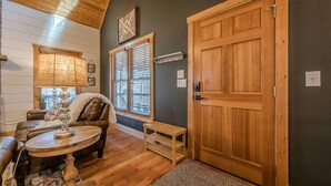 A cozy room with a wooden ceiling, a large wooden door, a cushioned seating area with a table, and a window with blinds. - Cozy Dahlonega Cabin w/ Hot Tub. Surrounded by wineries and 8 mins to downtown! (Lumpkin County)