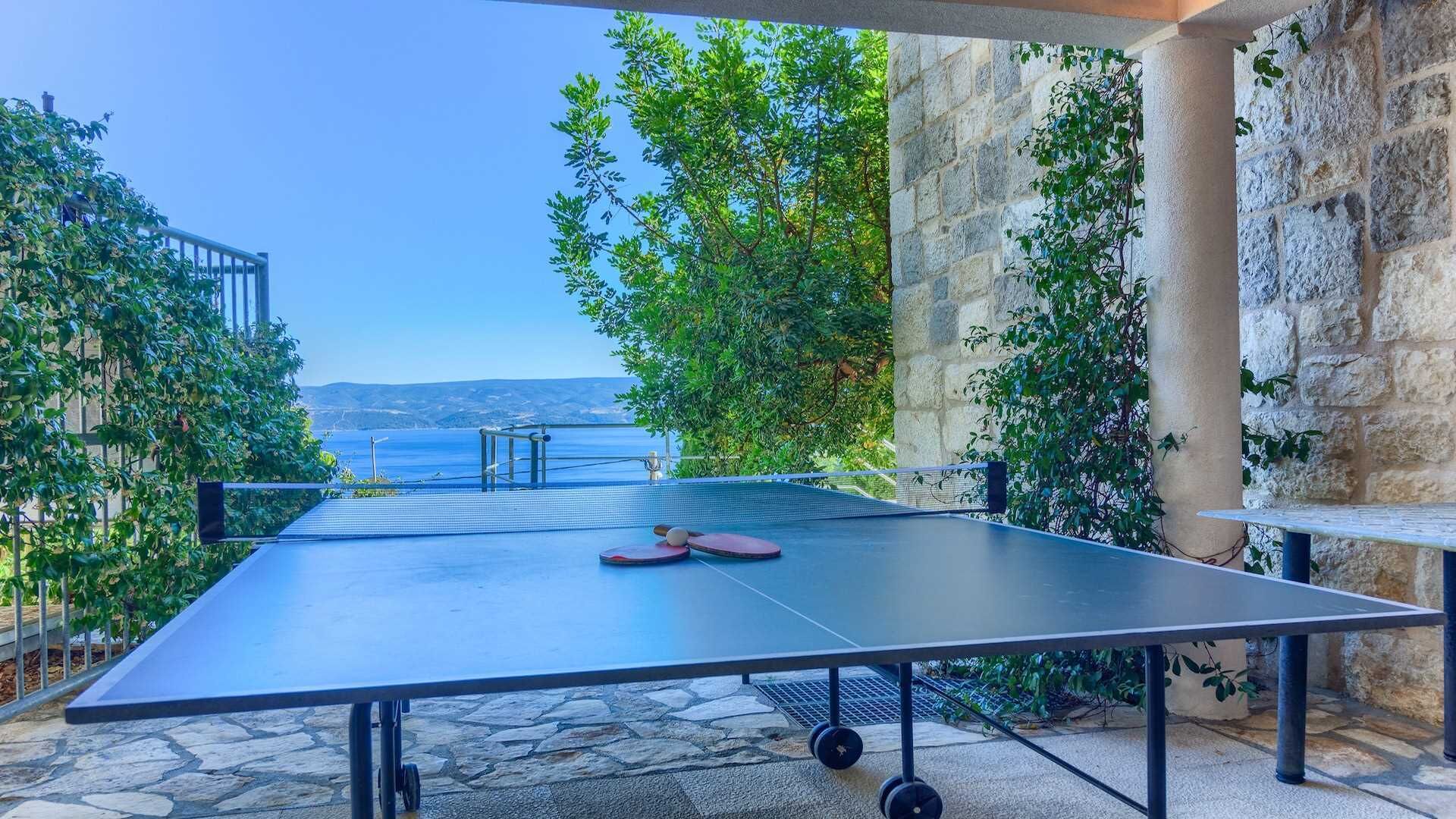 An outdoor ping pong table with a stone floor, surrounded by greenery and a stone wall, overlooking a body of water.