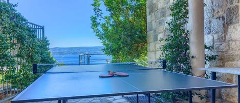 An outdoor ping pong table with a stone floor, surrounded by greenery and a stone wall, overlooking a body of water.