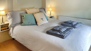 A neatly made bed with pillows, a folded blanket, and a stack of towels. There is a bedside table with a lamp and a book.