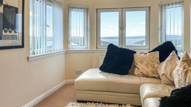 A corner living room with a beige leather sofa, a fluffy white rug, and a view of a body of water through large windows.
