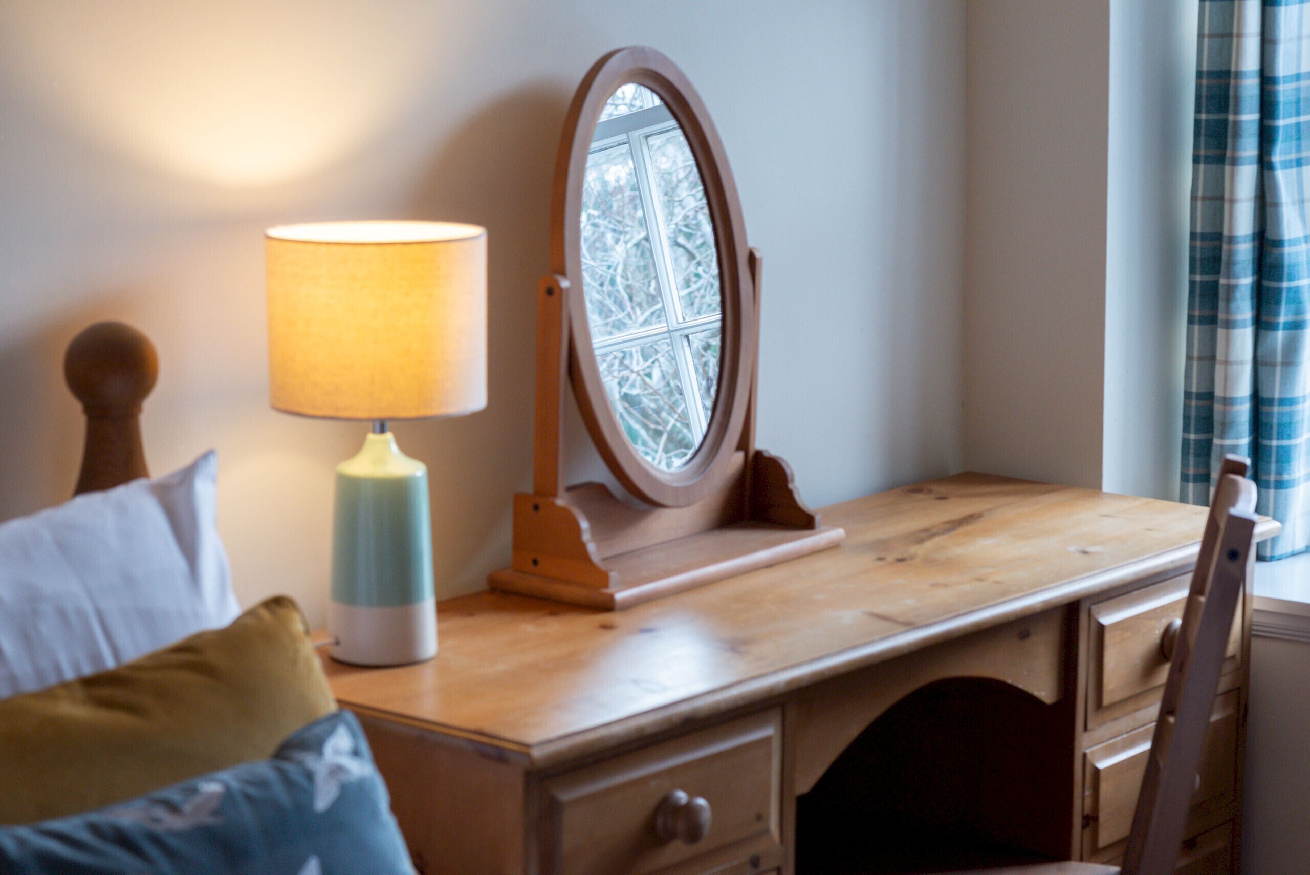 A wooden desk with a lamp, a mirror, and a window with a view of trees.
