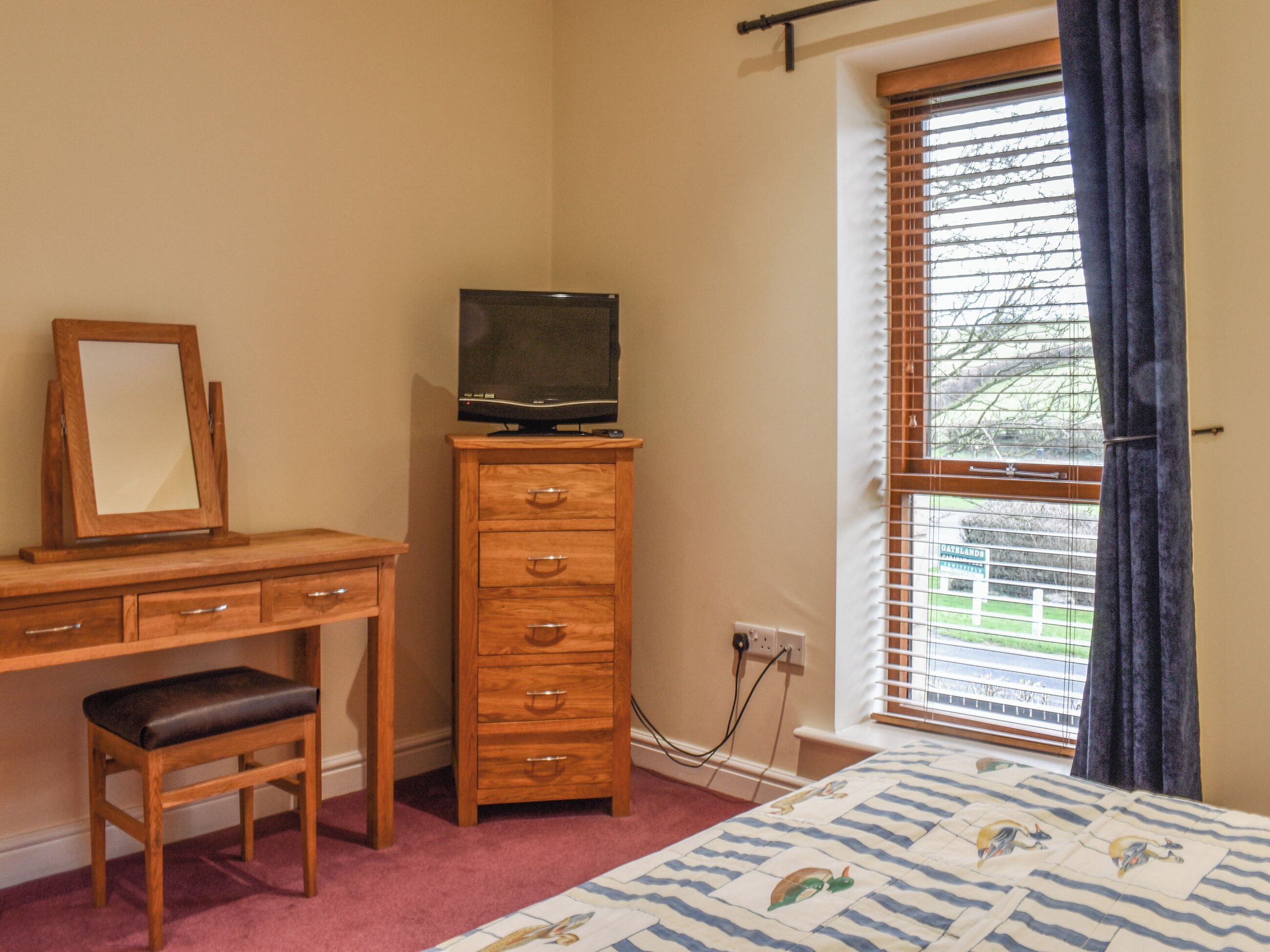 A bedroom with a wooden desk, a chair, a television on a dresser, a window with blinds, and a bed with a patterned bedspread.