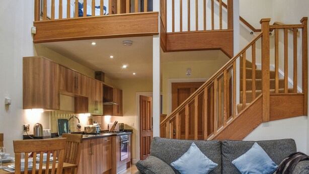 A spacious living area with a high ceiling, wooden staircase, and a kitchen with wooden cabinets and a dining table.