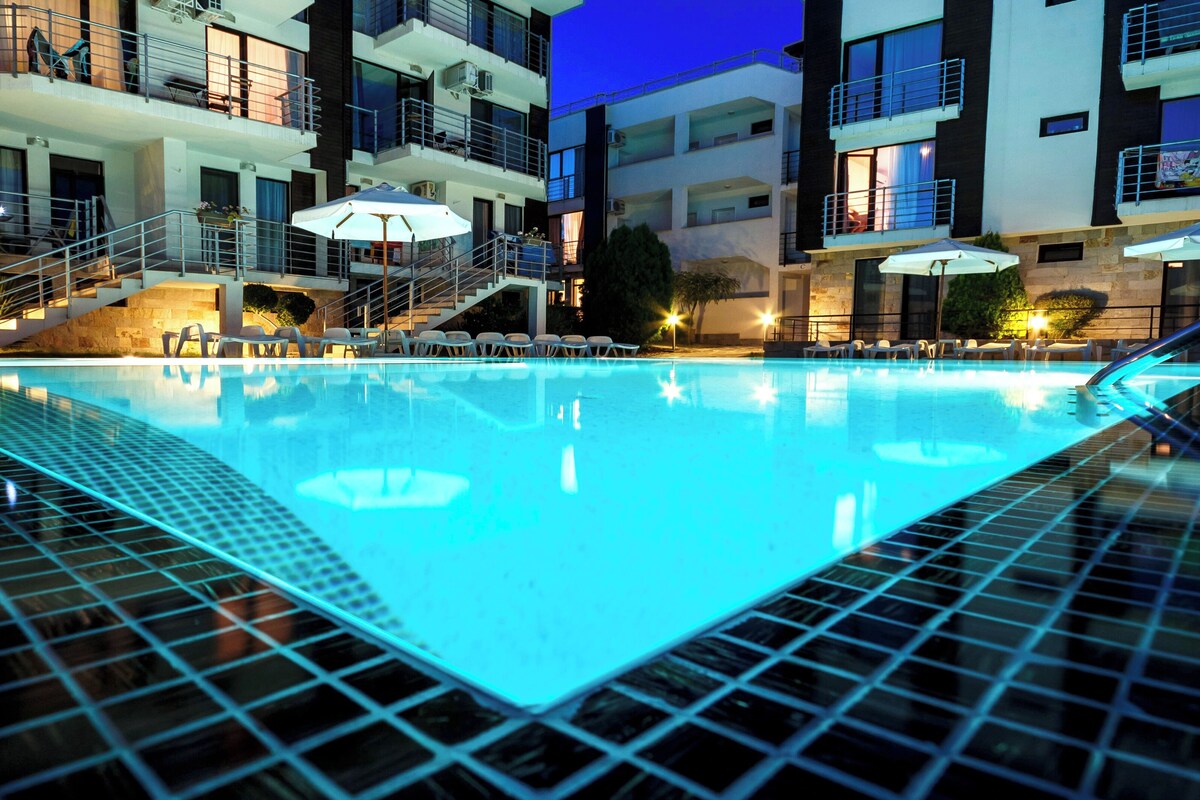 En swimmingpool med ett kaklat poolområde, omgiven av solstolar och parasoller, och intilliggande en flervåningsbyggnad med balkonger och utomhusmöbler.