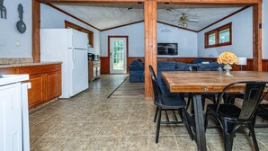 Eine geräumige Küche mit Kühlschrank, Herd und Holzschränken. Es gibt einen Essbereich mit Tisch und Stühlen, und im Hintergrund steht ein Sofa.