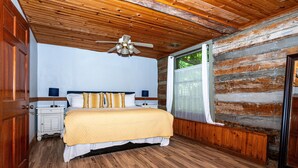 Ein Schlafzimmer mit Holzbalkendecke, einem Bett mit gelber Decke, einem Nachttisch und einem Fenster mit Blick ins Grüne.