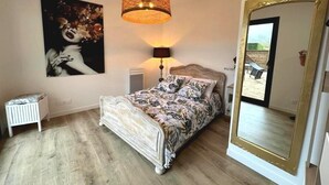 Une chambre à coucher comprenant un lit, une table de chevet, une lampe, un grand miroir et un mur orné d’un tableau encadré.