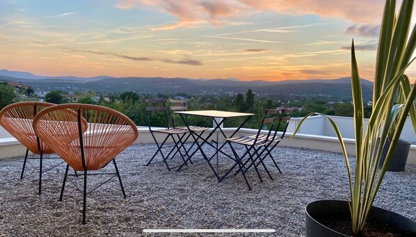 Una terraza con sillas de mimbre y una mesa, con vistas a un paisaje pintoresco con montañas y un atardecer.