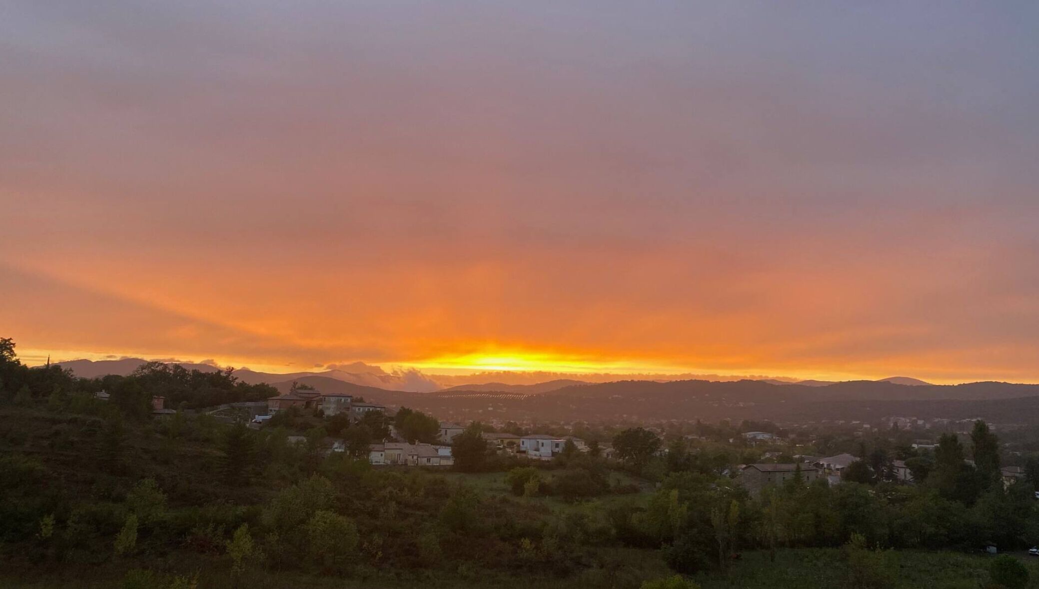 A landscape with a sunset over a town, featuring rolling hills and scattered buildings.