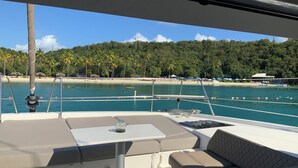 Cubierta de barco con asientos y una mesa, con vista a una playa con palmeras y un cielo azul despejado.