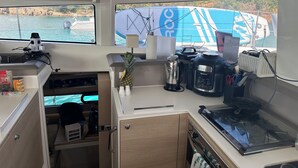 Cocina pequeña con estufa, cafetera y un refrigerador. Una ventana permite ver un barco y el agua.