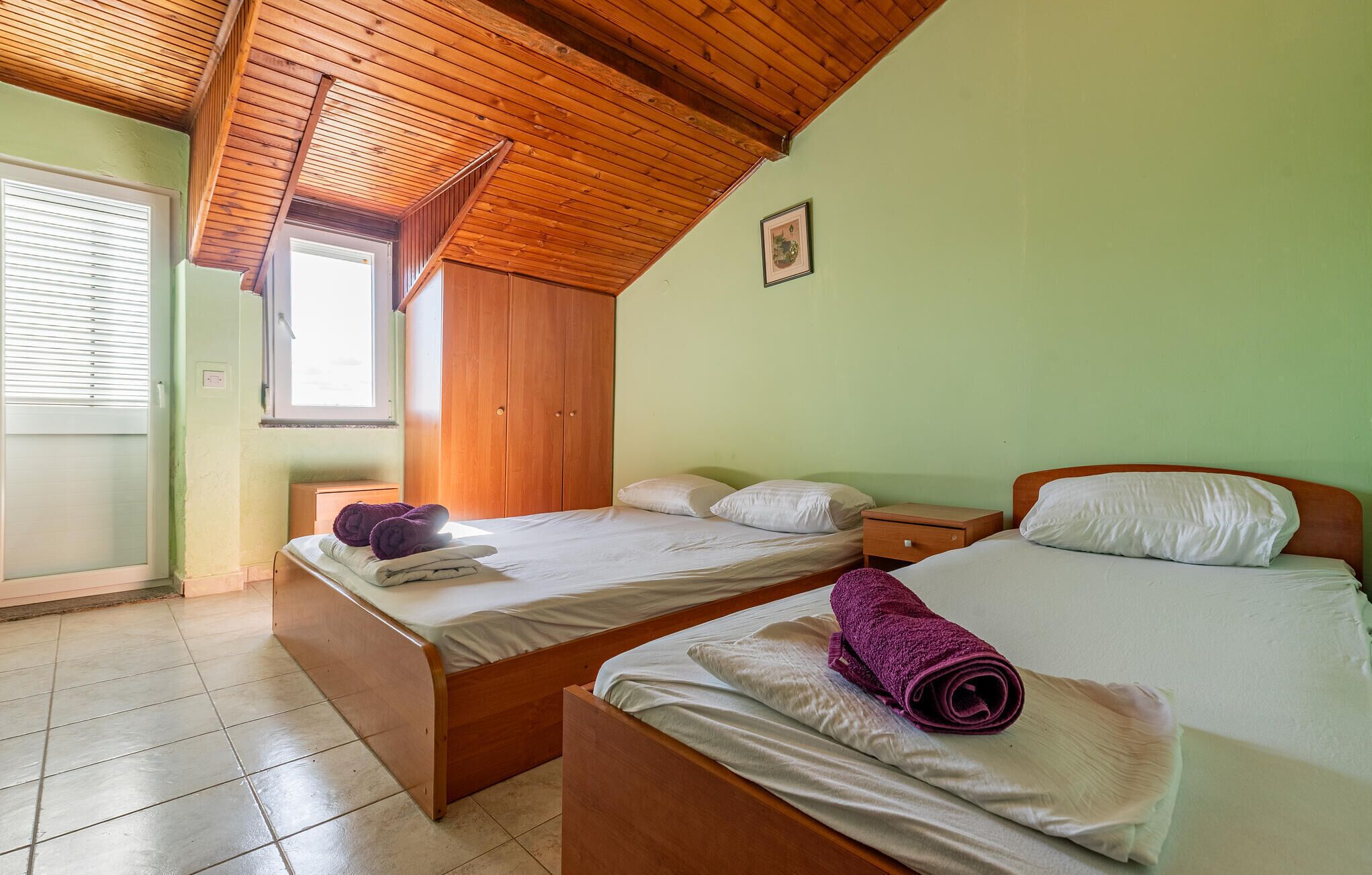 Ein Zimmer mit zwei Betten, Holzbalkendecke, einem Fenster mit Jalousien und einem kleinen Bild an der Wand.