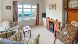 Una sala de estar con chimenea, un ventanal con cortinas, un sofá, un sillón y un reloj en la pared.