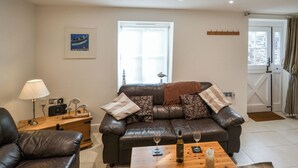 A living room with a leather sofa, a wooden coffee table with wine glasses and a candle, a lamp, and a framed picture on the wall. - 2 bedroom accommodation in Mawgan Porth (Mawgan Porth)