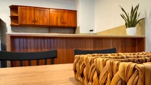 Een eetgedeelte met een houten tafel en stoelen, een geweven tafelkleed, houten kasten en een potplant.