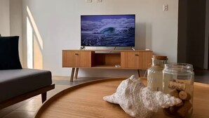 Een woonkamer met een televisie op een houten standaard, een bank en een ronde houten tafel met potten en een decoratief object.