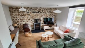 A living room with a stone wall, a fireplace, a television, a green sofa, a white sofa, and a dining area with a table and chairs. - TatîeMamîe, gîte Tata Jacqueline : grand gîte rénové proche mer, Bretagne (Plouha)