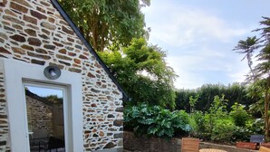 A stone building with a patio area featuring metal chairs and a table, surrounded by greenery. - TatîeMamîe, gîte Tata Jacqueline : grand gîte rénové proche mer, Bretagne (Plouha)