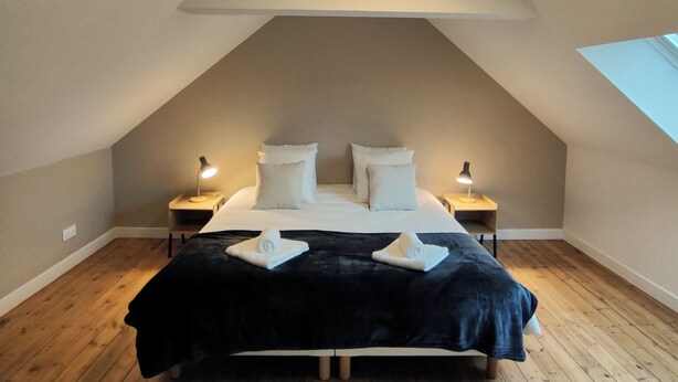 A bedroom with a sloped ceiling, a bed with white linens, two bedside lamps, and a large window. - TatîeMamîe, gîte Tata Jacqueline : grand gîte rénové proche mer, Bretagne (Plouha)