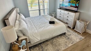 Un dormitorio con cama, televisión, cómoda y ventana que ofrece vistas a palmeras y una piscina.