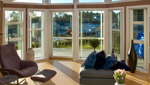 A sunlit room with a large window, a sofa with cushions, a chair, and a vase of flowers.