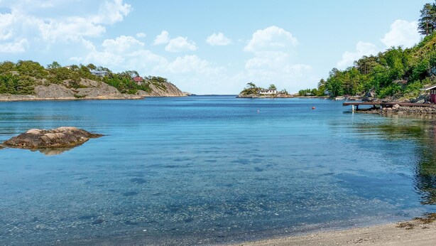A clear coastal view with calm waters, a sandy beach, and a small island with buildings.