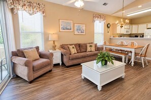 A living room with two sofas, a coffee table, a dining table with chairs, and a kitchen area in the background.