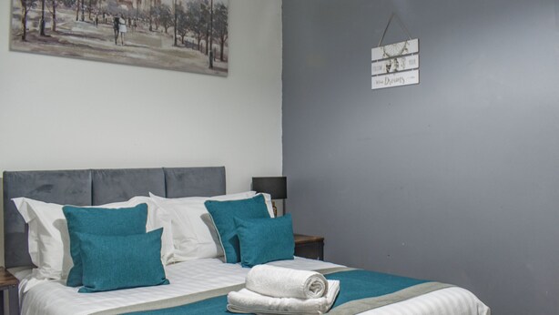 Un lit bien fait, recouvert d’une couvre-lit bleue et blanche, avec deux oreillers et une serviette enroulée. Un tableau est accroché au mur, et une pancarte avec l’inscription « SUIVEZ VOS RÊVES » est placée au-dessus du lit.