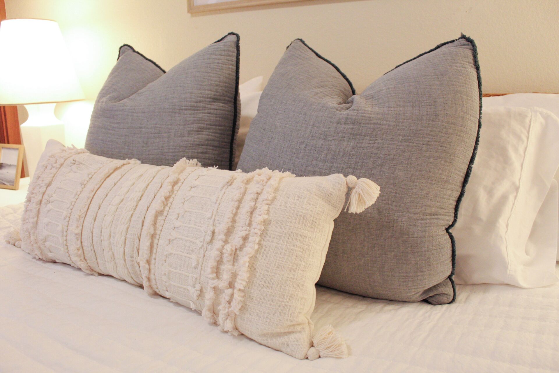 Un lit avec trois oreillers, un blanc et deux gris, recouvert d’une couverture blanche à texture.