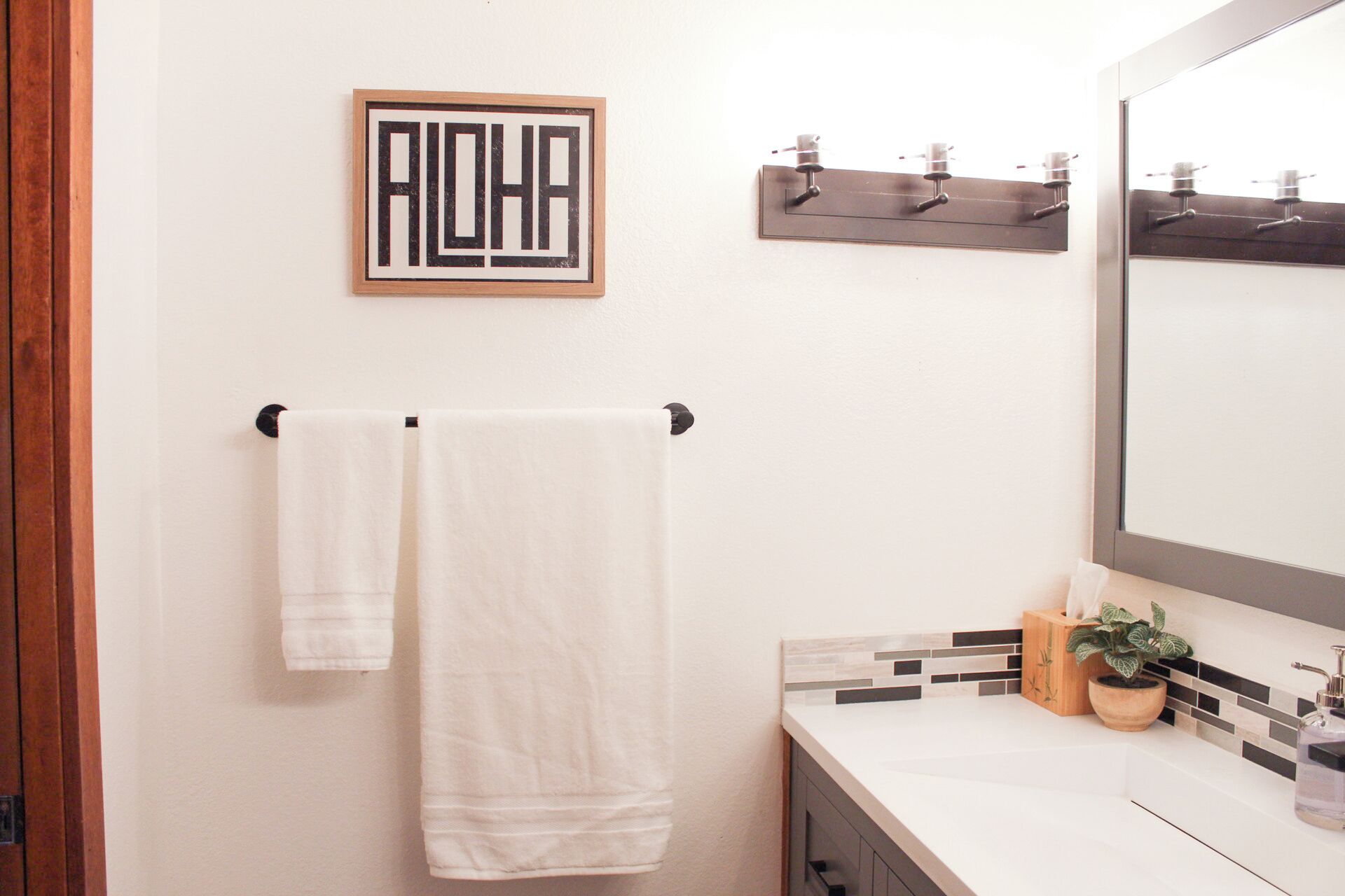 Une salle de bain avec deux serviettes blanches suspendues à une barre à serviettes noire, un panneau encadré avec le mot « ALOHA » accroché au mur, un miroir avec un cadre en bois et une petite plante en pot posée sur le comptoir.