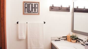 Une salle de bain avec deux serviettes blanches suspendues à une barre à serviettes noire, un panneau encadré avec le mot « ALOHA » accroché au mur, un miroir avec un cadre en bois et une petite plante en pot posée sur le comptoir.